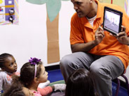 A teacher sits on a chair in front of students seated on the floor. He is showing something on an iPad.