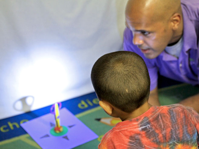 A teacher and a student are looking at a spot lit wall, in front of which is a rotation mat, with a pair of scissors standing upright in the center of the mat, stabilized in Play-Doh.