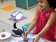 A student holds a paper plate, on which sits a shadow sculpture, made of a piece of Play-Doh with popsicle sticks and pipe cleaners sticking out of it. A lamp sits on the table next to her.