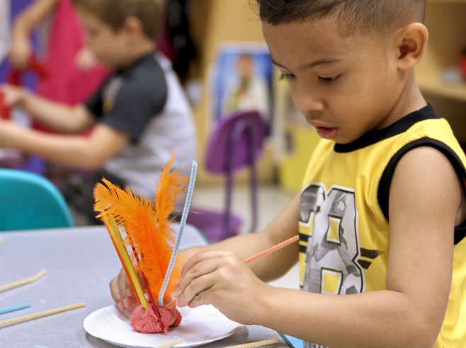 A student holds a paper plate, on which sits a shadow sculpture, made from Play-Doh that has popsicle sticks and pipe cleaners sticking out of it.