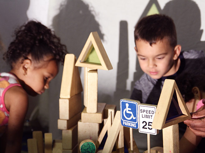 Two students are building a structure, using wooden blocks of different shapes. A light casts shadows of the structure on the wall behind them.