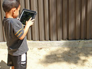 A student holds an iPad up in a sandy playground. He is taking a picture of a shadow made by a tree.
