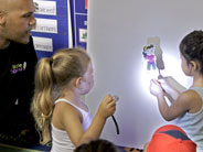 One student holds a hand-made shadow puppet against a piece of white poster-board on the wall. Another student shines a flashlight at it, creating a shadow.
