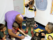 Students on the floor crowd around a piece of paper. One student stands, leaning over the paper and shining a flashlight on it while a teacher draws with a marker on the paper.