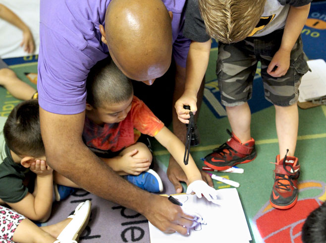 Students on the floor crowd around a piece of paper. One student shines a flashlight over a student's hand above the paper while a teacher draws with a marker on the paper.