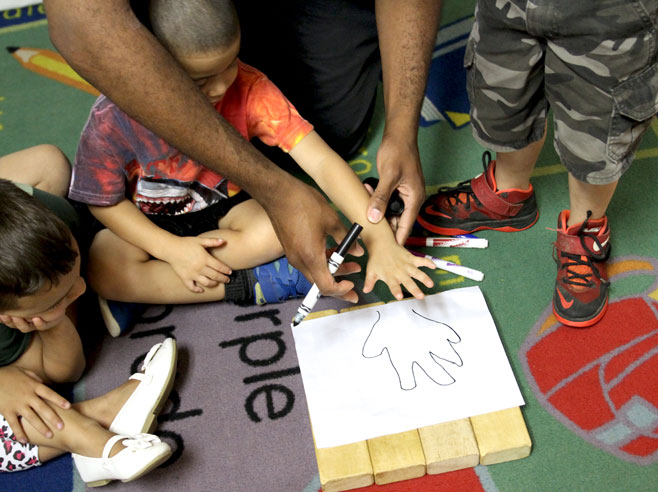 Students gather around a piece of paper on the floor as a teacher lifts up a student's hand. The teacher has just finished tracing the student's hand on the piece of paper.