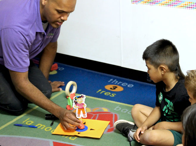 A teacher is placing a hand-made shadow puppet on the blue circle, in the middle of a rotation mat, laid on the floor. The craft stick on the puppet is stuck in Play-Doh, so that the shadow puppet stands up.