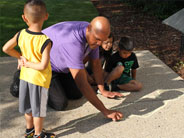 Outside, a student stands, while a teacher traces his shadow with chalk on the sidewalk.
