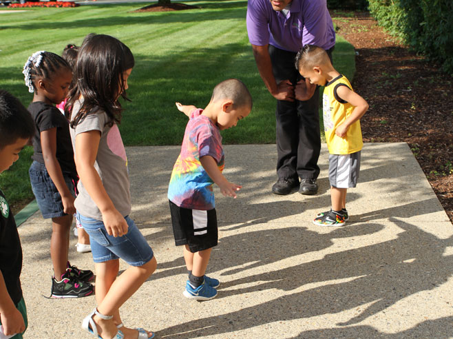 Outdoors, a teacher can be seen bending to talk to a group of students, who play with their shadows on the sidewalk.
