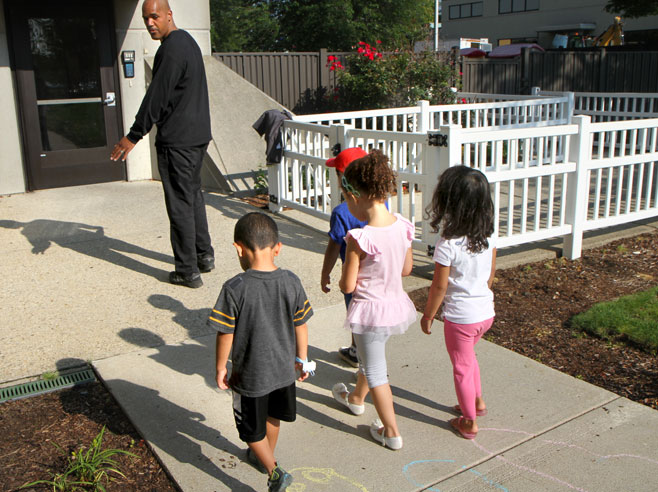 Four students walk behind a teacher, towards the door of a building. It is sunny, and their shadows are clearly visible.