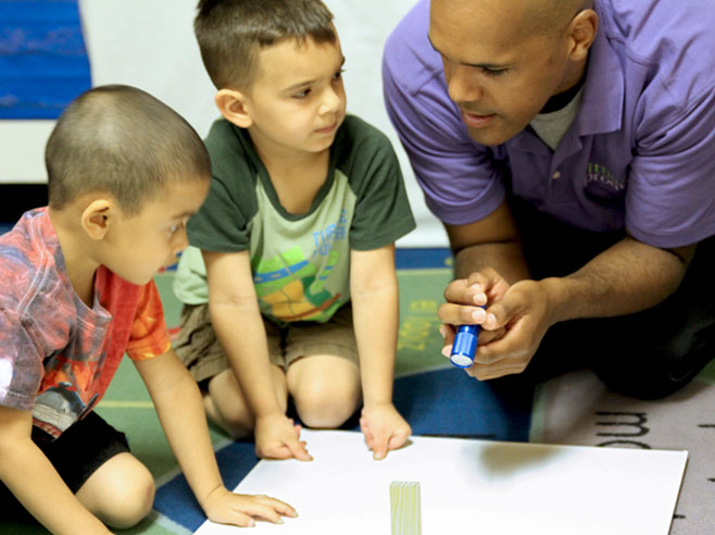 A piece of white poster-board lies on the floor with a wooden block on top. A teacher shines a flashlight at the block while students listen to the teacher.