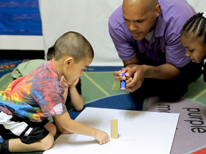 A piece of white poster-board lies on the floor with a wooden block on top. A teacher shines a flashlight at the block, and one student places a craft stick by the block.