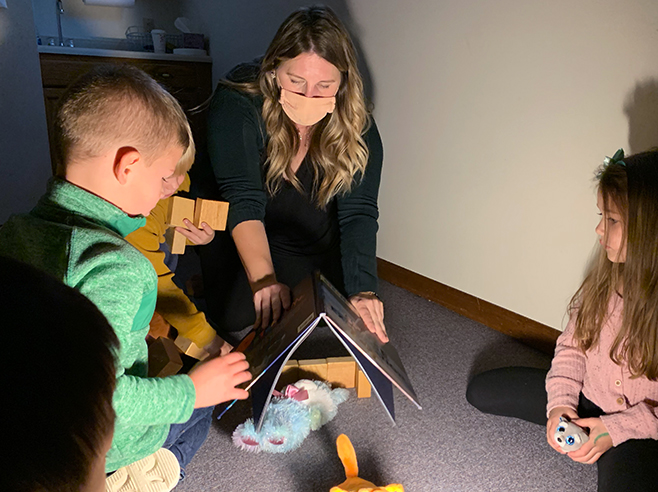 Light shining on teacher covering stuffed animal with a book. Two students watching.